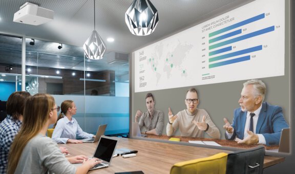 Vier Personen sitzen an einem Tisch und sind im hybriden Microsoft Teams FrontRow Meeting mit drei weiteren virtuell zugeschalteten Personen