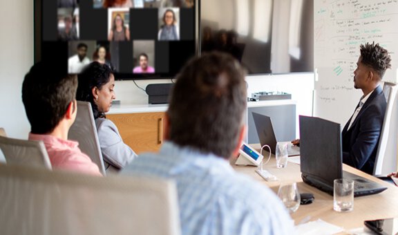 Sechs Personen sitzen in einer Videokonferenz an einem Tisch mit eigenen Laptops und Tablets, schauen auf einen Bildschirm