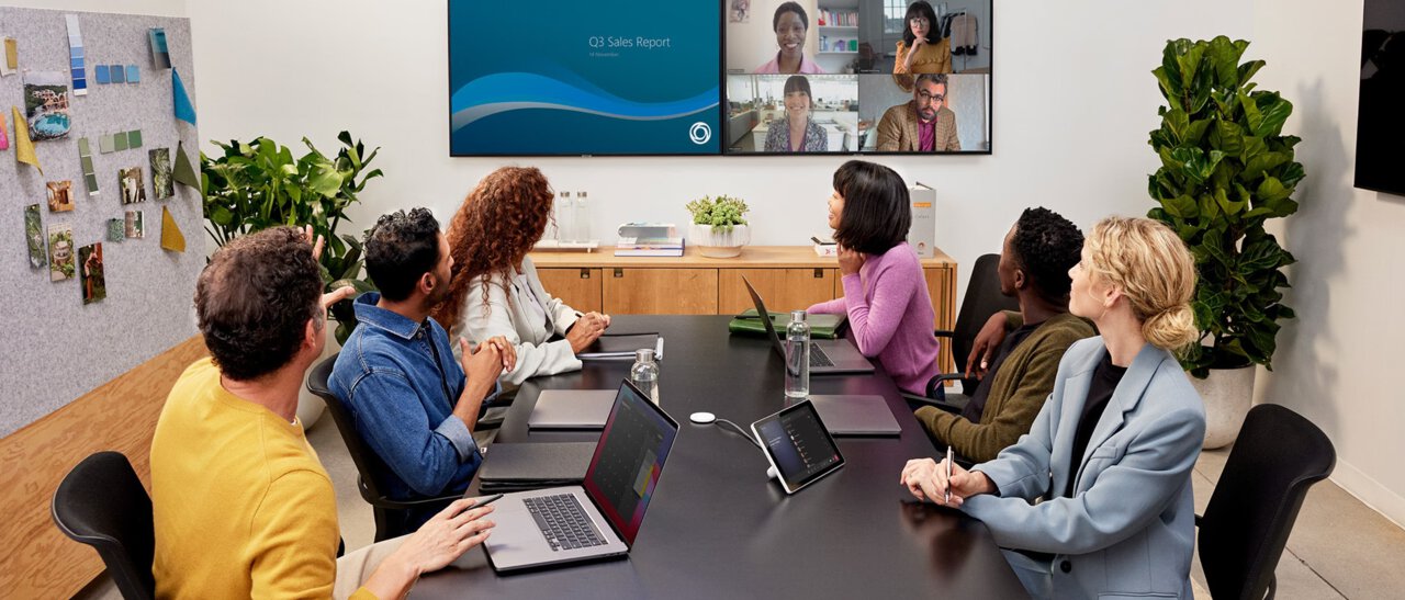Sechs Personen sitzen an einem Tisch mit mehreren Laptops und schauen auf einen Doppelbildschirm mit einer Cisco Webex Room Bar