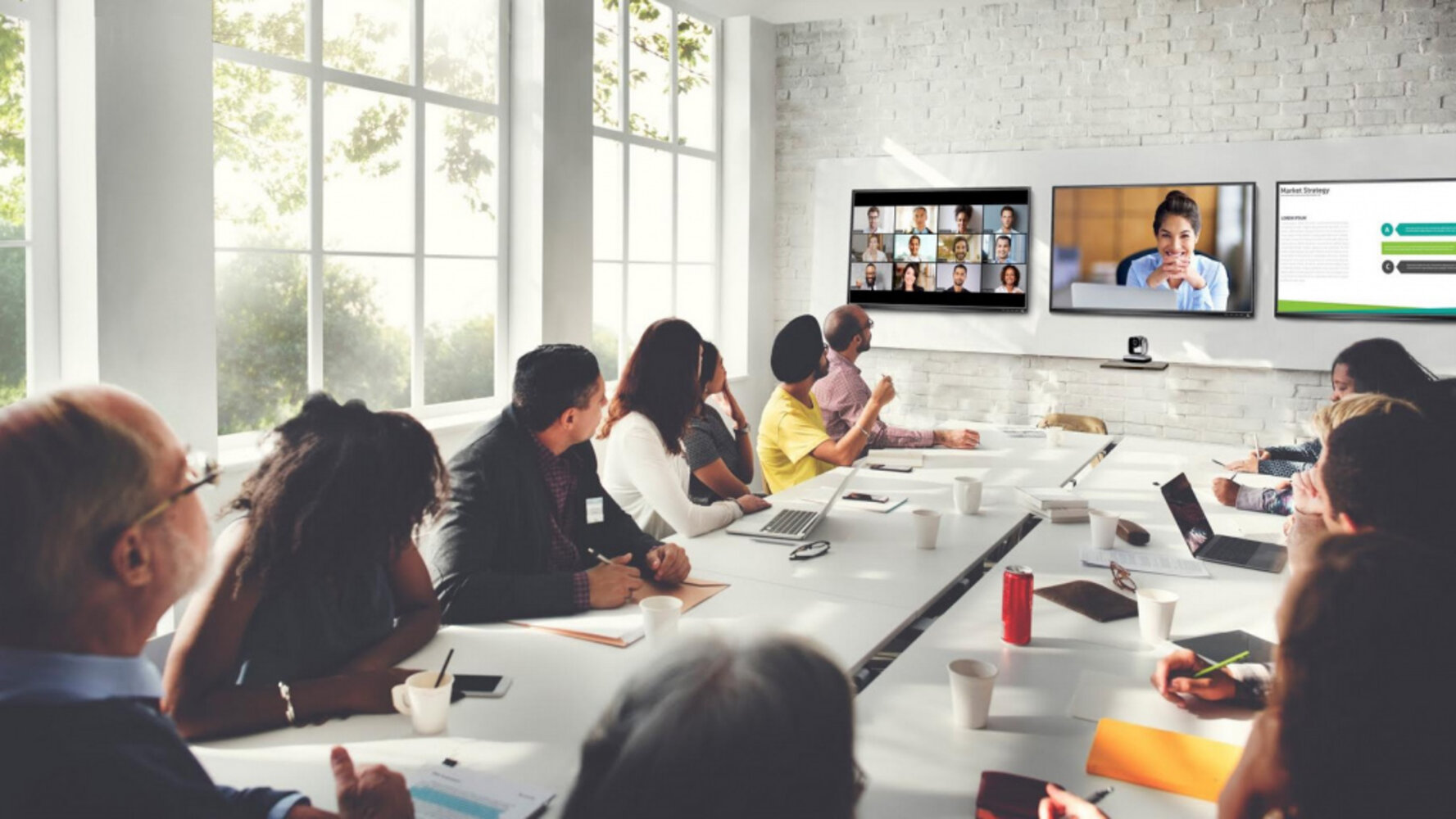 Viele Menschen in einem Konferenzraum, sitzen an einem Tisch und halten auf drei Bildschirmen ein Webinar, Linksseitig sind die anderen Webinarteilnehmer sichtbar, mittig die referierende Person und auf dem rechten Bildschirm die Inhalte