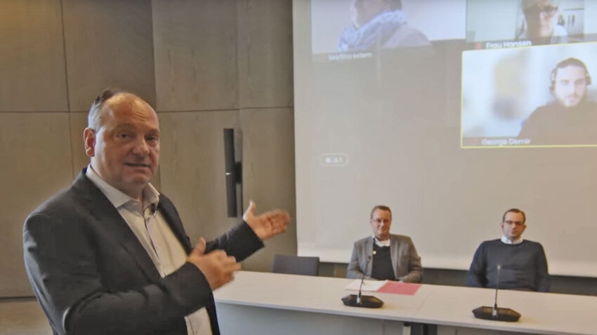 Ein mann der in einem Sitzungssaal auf das podium zeigt, an dem Menschen sitzen, im Hintergrund werden die hybriden Teilnehmer per Beamer an die Wand gestrahlt