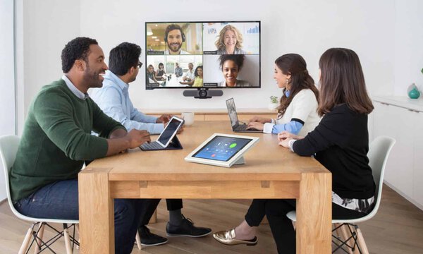 Vier Personen sitzen in einem Konferenzraum in einem hybriden Zoom-Meeting, schauen auf einen Bildschirm mit den anderen Meetingteilnehmern