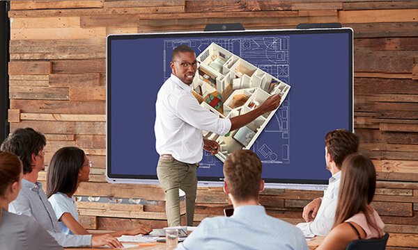 Sieben Personen in einem modernen Meetingraum, eine Person vorn zeigt auf einem interaktiven Whiteboard ein 3D Modell eines Gebäudes und spricht, 