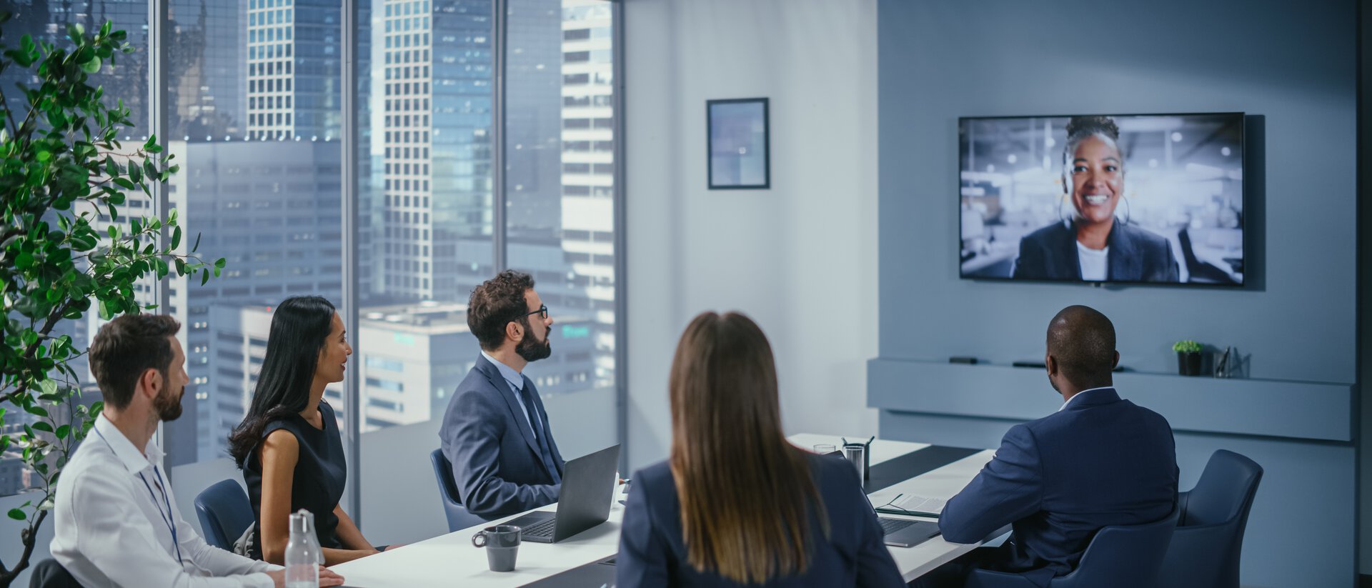 Fünf Personen sitzen im Meetingraum an einem Tisch und halten eine Videokonferenz mit einer weiteren Person ab, eleganter, moderner Konferenzraum 
