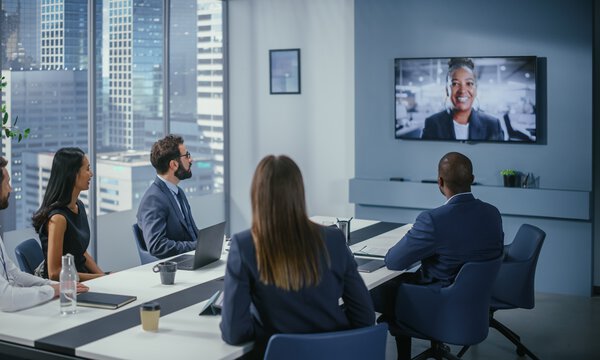 Fünf Personen sitzen im Meetingraum an einem Tisch und halten eine Videokonferenz mit einer weiteren Person ab, eleganter, moderner Konferenzraum 