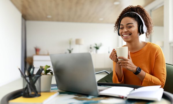 Junge Frau vor dem Laptop in einem Videocall, hält eine Tasse und hat ein Headset auf