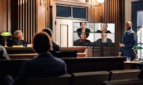 Mehrere Personen in einem Gerichtssaal schauen auf einen Bildschirm mit weiteren Zeugen und Richtern