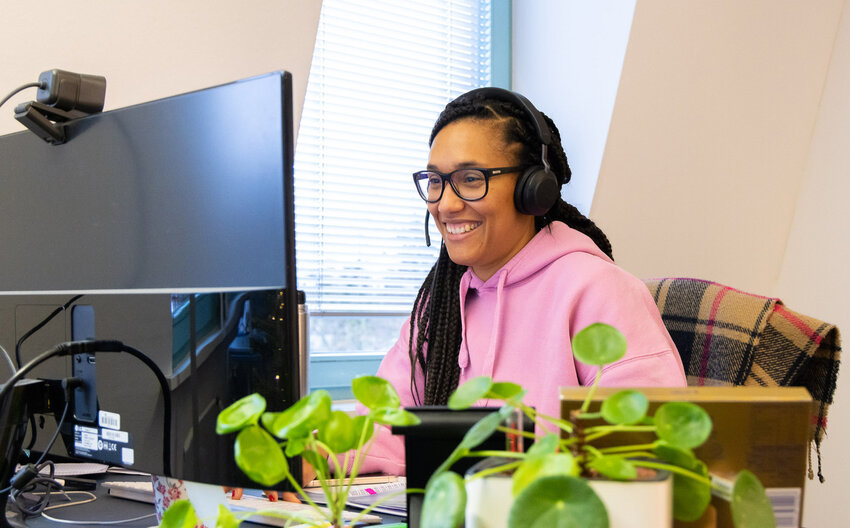 Frau am Arbeitsplatz mit einem Großen Monitor, Webcam, Headset und Pflanzen auf dem Tisch