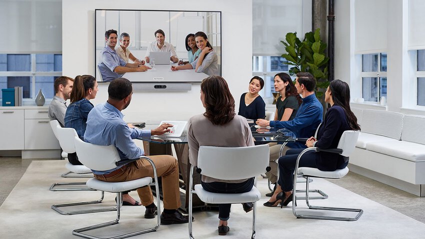 Acht Personen in einem mittelgroßen Meetingroom im Cisco Webex Videocall, stirnseitig ein großes Display mit den weiteren Meetingteilnehmern, darunter eine Videobar