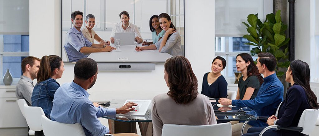 Acht Personen in einem mittelgroßen Meetingroom im Cisco Webex Videocall, stirnseitig ein großes Display mit den weiteren Meetingteilnehmern, darunter eine Videobar