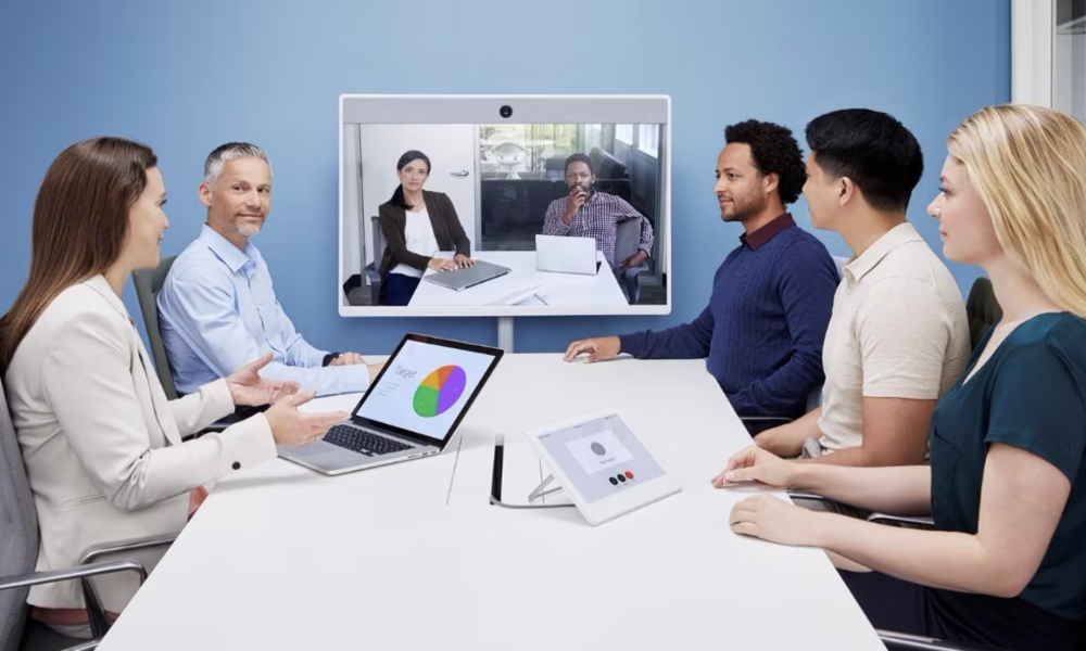 Fünf Personen in einem mittelgroßen Meetingraum in einem hybriden Meeting mit zwei weiteren, die auf dem Bildschirm sichtbar sind, auf dem Konferenztisch stehen eine Raumsteuerung und ein Laptop