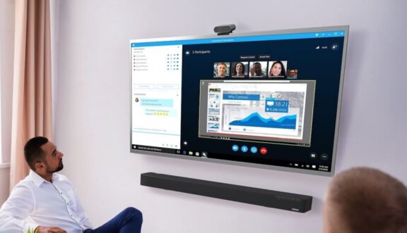Zwei Männer sitzen vor einem Display, auf dem eine Videokonferenz läuft, darunter ist die Lenovo ThinkSmart Bar installiert