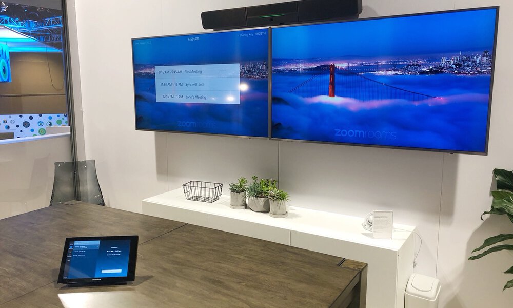 Blick in einen mittelgroßen Konferenzraum mit dualem Bildschirm und einem Touchpad zur Steuerung, auf den Displays ist der Startbildschirm von Zoom sichtbar 