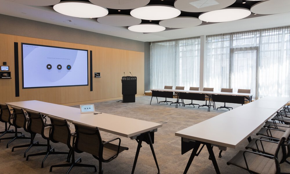 Ein leerer Konferenzraum mit einem großen Display in der Wand, vor dem drei längliche Tische stehen die mit der Wand ein Viereck bilden