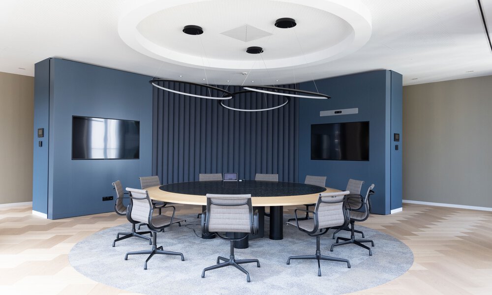 Ein leerer, runder Konferenzraum mit zwei Displays in den Wänden und einem runden Tisch in der Mitte, um den herum Stühle stehen